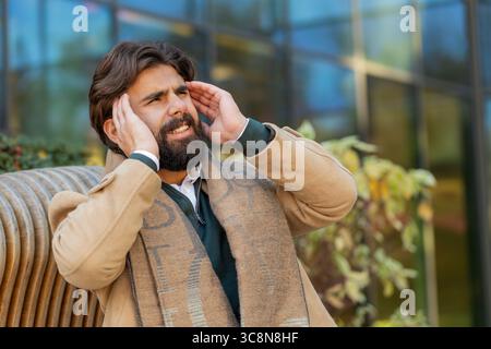 Der kranke arabische Indianer hat Kopfschmerzen auf der Stadtstraße. Der junge Tourist massiert ihre Stirn und Schläfen mit den Händen gegen das Gebäude. Verärgerter hinduistischer Geschäftsmann, der an Migräne leidet. Stockfoto