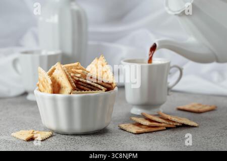 Knusprige quadratische Cracker und schwarzer Kaffee. Im Hintergrund wird schwarzer Kaffee aus einer Kaffeekanne in eine Tasse gegossen. Stockfoto
