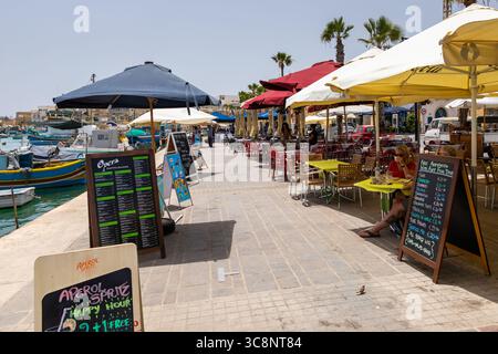 Marsaxlokk, Malta – 22. Juni 2021: Cafés und Restaurants mit Sitzgelegenheiten im Freien und farbenfrohen Sonnenschirmen entlang der Hafenpromenade. Stockfoto