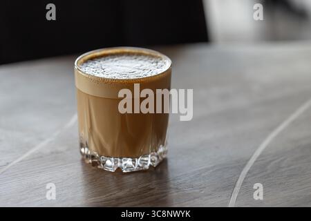 Flach weiß. Ein warmes, schaumiges Kaffeegetränk, serviert in einem klaren Glas auf einer rustikalen Tischplatte. Eine gemütliche und einladende Atmosphäre für die morgendliche Routine, entspannen Sie sich Stockfoto