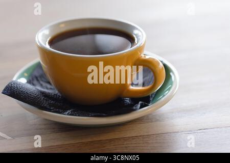 Eine hellgelbe Tasse gefüllt mit heißem schwarzem Kaffee auf einer Holzoberfläche. Die Szene weckt Wärme und eine gemütliche Atmosphäre, perfekt für morgendliche Routen Stockfoto