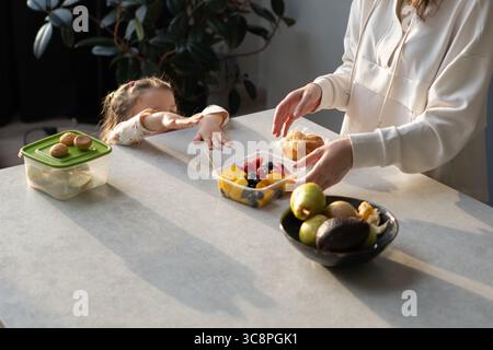 Ein Mädchen zu Hause, das zu einem Obstbehälter greift, eine Frau, die Essen auf den Tisch legt Stockfoto