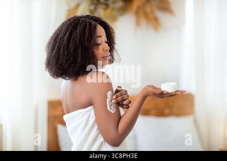 Schönheitsbehandlung für gesunde Haut. Wunderschöne schwarze Frau im Badetuch Anwendung Körpercreme oder Butter auf der Schulter zu Hause Stockfoto