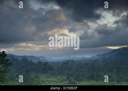 Eine kraftvolle und stimmungsvolle Szene entfaltet sich, während sich Sturmwolken um den fernen Gipfel des Mount Agung auf Bali winden. Nebelschichten driften über den Regenwald und Stockfoto
