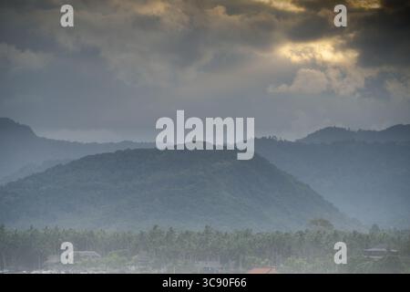 Eine ruhige Landschaft auf Bali entfaltet sich unter einem stimmungsvollen, bewölkten Himmel. Das Bild zeigt mehrere Schichten bewaldeter Hügel, die langsam in die Ferne versinken. Stockfoto
