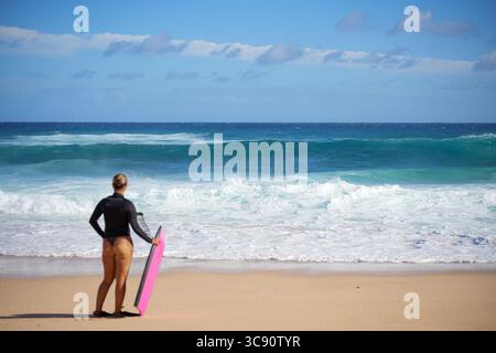 Bodyboarder steht am stürmischen Strand, hält das Bodyboard aufrecht im Sand und blickt auf die große Meeressturmwelle – zwischen Angst und Nervenkitzel, mit Blick auf das Meer Stockfoto