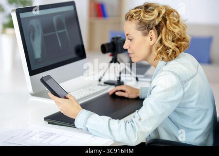 Eine junge, attraktive blonde Frau sitzt an einem modernen Schreibtisch mit einem Computermonitor vor sich, hält und schaut auf ein Smartphone. Sie scheint konzentriert zu sein Stockfoto
