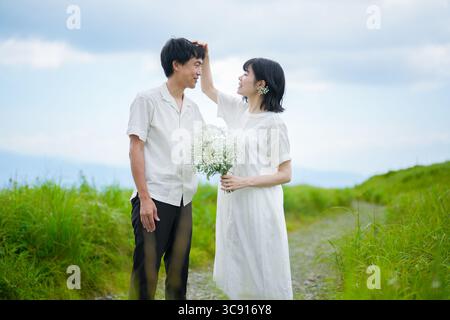 Ein Paar in ihren 30ern steht auf einem Bergweg, die Frau in einem weißen Kleid mit mittellangen Haaren hält Baby Atem an und fixiert das Haar des Mannes als Stockfoto