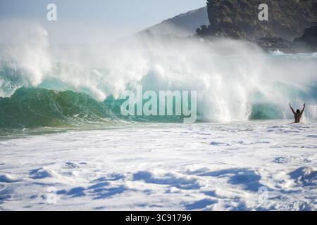 Der Alleinschwimmer hebt seine Arme im Ozean, während die gewaltige Welle herunterstürzt, die Wassernebel hoch fliegt und dramatische Klippen im Hintergrund den Nervenkitzel fängt Stockfoto