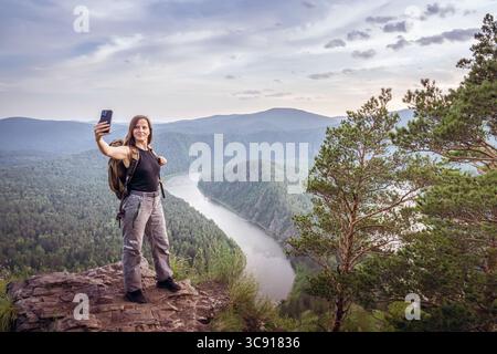 Ein Wanderer steht auf einem felsigen Felsvorsprung und macht ein Selfie vor einer atemberaubenden Kulisse aus üppig grünen Wäldern und einem sich windenden Fluss. Eine weibliche Alleinreise Stockfoto