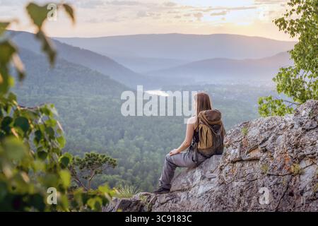 Eine junge Frau sitzt auf einem großen Felsen und blickt auf ein riesiges Tal, das von einem warmen Sonnenuntergang beleuchtet wird. Die Landschaft ist mit dichten Bäumen und einem sich windenden Fluss im Th Stockfoto