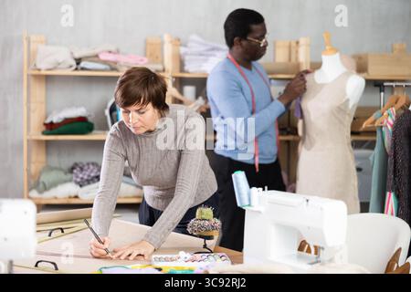 Frau zeichnet das Kleid des Produkts auf Papier. Begleitarbeit im Hintergrund mit Schaufensterpuppe Stockfoto