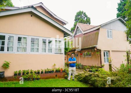 Mann steht vor einem traditionellen Landhaus mit weißen Fenstern und Garten in üppiger grüner Umgebung. Stockfoto