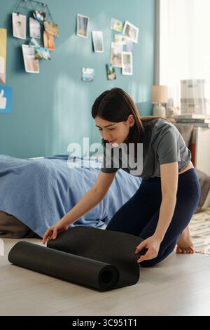 Junge Erwachsene kaukasische Frau kniet auf dem Boden und bereitet Yoga-Matte im Schlafzimmer vor, konzentriert sich auf das Abrollen der Matte mit beiden Händen, zwangloses Zuhause mit persönlichen Fotos an der Wand Stockfoto