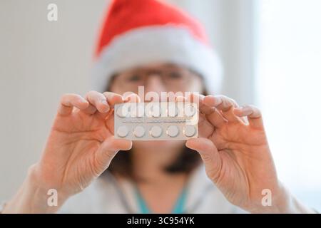 Lächelnde Ärztin in Weihnachtsmütze und weißer Uniform, die Arzneimitteltabletten hält. Festlicher Gesundheitsmoment, verschreiben Behandlung für Wintergrippe oder Kälte während der Weihnachtszeit. Fürsorgeexperte. Stockfoto