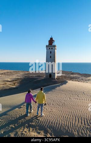 Ein Paar genießt einen romantischen Spaziergang auf dem goldenen Sand Dänemarks, Hand in Hand. Der historische Leuchtturm steht an einem sonnigen Tag vor der kristallklaren Kulisse des Ozeans. Rubjerg Knude Stockfoto
