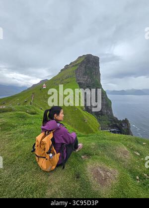 Ein Reisender sitzt friedlich auf einem grasbewachsenen Hügel mit Blick auf die atemberaubenden Klippen der Färöer Inseln. Asiatische Frau besucht Kallur Lighthouse Kalsoy Island Stockfoto