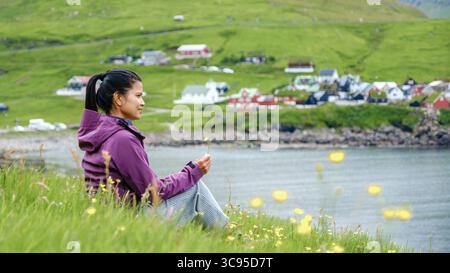 Eine Frau sitzt friedlich auf einem grasbewachsenen Hügel auf den Färöern, umgeben von lebendigen Wildblumen. Elduvik Village Stockfoto