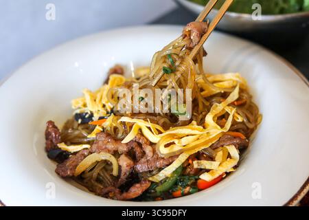 Ein paar Essstäbchen heben eine Portion Rindfleisch Japchae. Dieses klassische Gericht kombiniert kauende Glasnudeln, zartes Rindfleisch und Gemüse. Stockfoto