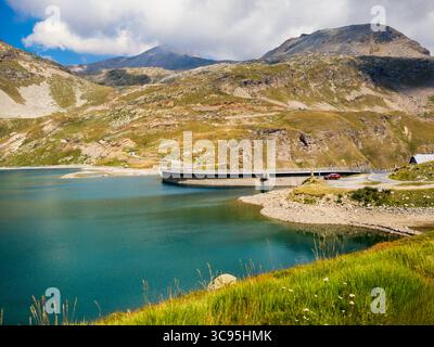 Ein malerischer Blick auf einen kleinen alpinen Agnel-See, der von felsigen Küsten und grünen Hängen umgeben ist, als Berge unter bewölktem Himmel im Hintergrund in Italien. Stockfoto