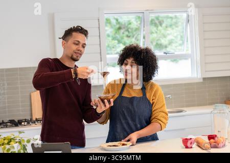 Afroamerikanisches Paar in Schürzen, das Schokoladensoße in eine Glasschale auf der Marmor-Kücheninsel löffelt Stockfoto