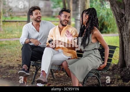 Eine fröhliche und abwechslungsreiche Gruppe von drei jungen Freunden, darunter eine stilvolle Schwarze, sitzt auf einer Parkbank, lacht und genießt eine positive Co Stockfoto