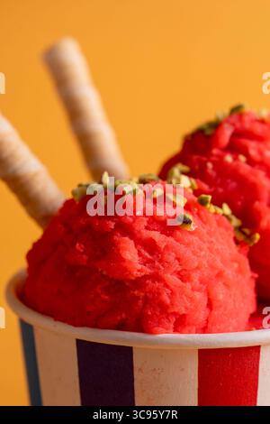 Ein köstliches Makrofoto von handwerklichem Erdbeersorbet-Schaufeln in einem Pappbecher, verziert mit Pistazien und Waffeln auf einem hellen orangefarbenen B Stockfoto