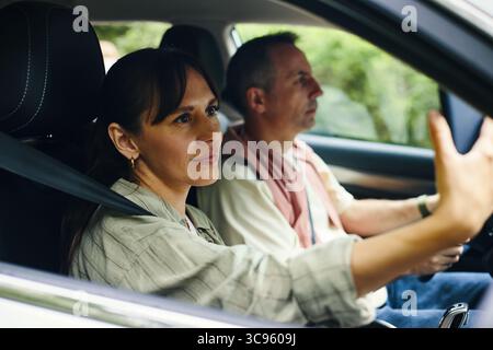 Kaukasische Frau mittleren Alters, die auf dem Beifahrersitz sitzt und Selfie mit dem Smartphone macht, während kaukasischer Mann mittleren Alters während einer Familienreise durch die Landschaft Auto fährt Stockfoto