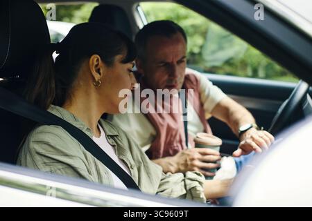 Kaukasische Frau mittleren Alters, die auf dem Beifahrersitz sitzt und mit einem kaukasischen Mann mittleren Alters spricht, der während eines Familienausflugs Auto fährt, Mann mit einer Kaffeetasse, beide mit Sicherheitsgurten Stockfoto