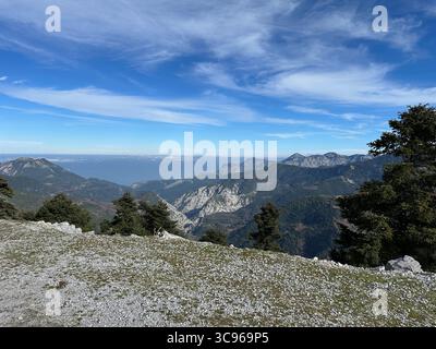 Weitläufige Berglandschaft mit geschichteten Bergrücken, Kiefernwäldern und einem Fernblick auf das Meer unter einem leuchtend blauen Himmel mit hellen Wolkenformationen. Stockfoto