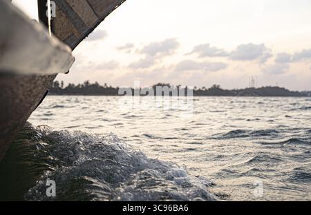 Blick auf das verwitterte Holz des Bootes, das durch das türkisfarbene Wasser in Richtung der fernen, trüben Küste unter hellem Himmel, Lamu, Kenia, schneidet. Stockfoto