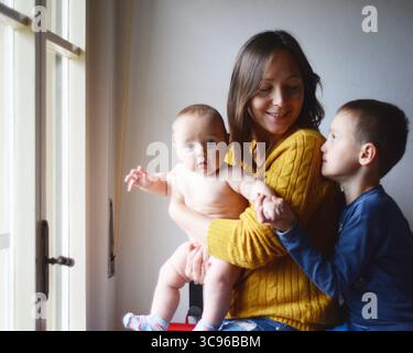 Die Mutter hält ihr Baby, lächelt warm, während sie mit einem älteren Kind am Fenster sitzt. Die Szene fängt einen Moment der Zärtlichkeit und Verbindung ein Stockfoto