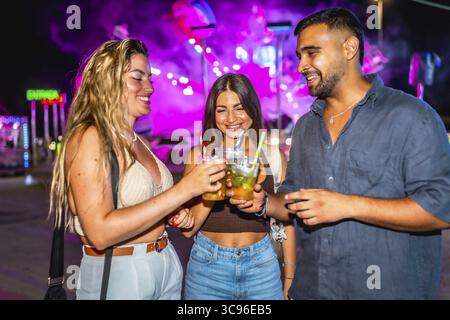 Drei glückliche Freunde, die abends im Vergnügungspark alkoholfreie Getränke tosten und gemeinsam Spaß haben Stockfoto