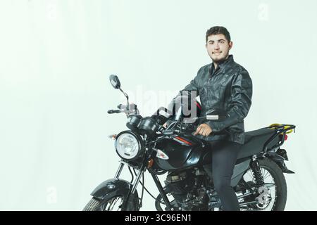 Lächelnder Motorradfahrer, der auf seinem Motorrad sitzt und einen Helm hält, isoliert. Porträt eines gutaussehenden Bikers auf Motorrad auf isoliertem Hintergrund Stockfoto