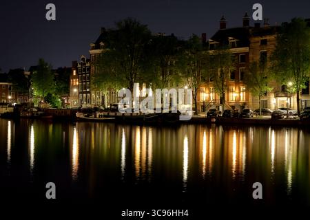 Glitzernde Hausboote und historische Fassaden säumen den Amstel-Kanal in Amsterdam, deren warme Lichter nachts in einer ruhigen, lang belichteten Reflexion tanzen Stockfoto
