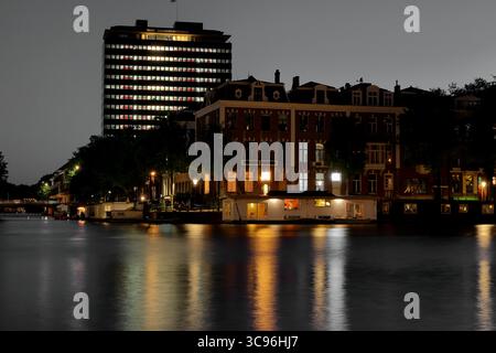 Historische Kanalhäuser und der beleuchtete DNB-Turm schimmern am ruhigen Amsterdamer Kanal in einer stimmungsvollen, lang ausgesetzten Nachtlandschaft und erinnern an urbane Ruhe Stockfoto