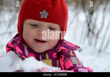 Ein Kind mit rotem Hut und farbenfrohem Mantel spielt gerne im Schnee. Der fröhliche Ausdruck fängt die Freude des Winters ein. Schneeflocken Stockfoto