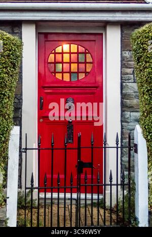 Eine rote Tür mit einem Buntglasfenster ist der Mittelpunkt dieses Bildes. Die Tür ist von weißen Zierleisten eingerahmt und von grünen Hecken flankiert. Eine schwarze Schmiede Stockfoto
