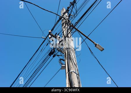 Ein Betonmast mit Stromleitungen und Kabeln vor einem hellblauen Himmel, der ein komplexes Netz von Infrastrukturen schafft. Stockfoto