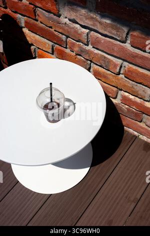 Ein erfrischender Eiskaffee steht auf einem weißen Tisch mit rustikalem Backsteinboden. Die Komposition hebt den Kontrast und die Details hervor. Stockfoto