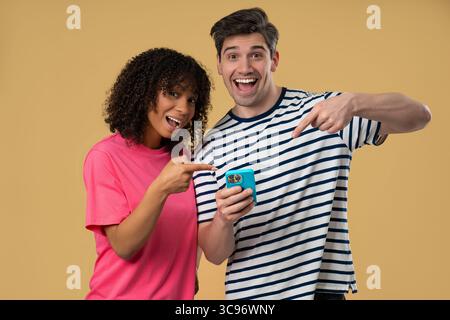 Interrassisches Paar zeigt auf Mobile. Frau Mann lächelt mit Smartphone auf Beige Stockfoto