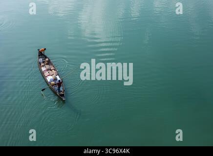Sunamganj, Bangladesch - 27. Januar 2019: Der Blick auf ein schlankes Boot gleitet über das ruhige, türkisfarbene Wasser, das seine Ladung und seine Besatzung unter dem riesigen Himmel trägt. Stockfoto