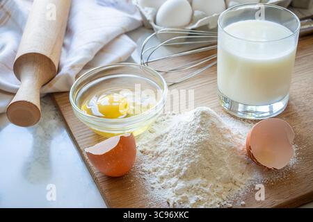 Mehl, ein Ei und Milch sind ordentlich auf einer Holzoberfläche angeordnet, mit einer Nudelnadel und Eierschalen in der Nähe zum Backen. Stockfoto