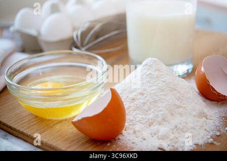 Mehl wird auf einem Holzbrett zusammen mit zerrissenen Eiern und einem Glas Milch aufgehäuft, bereit für Backzubereitungen in der Küche. Stockfoto