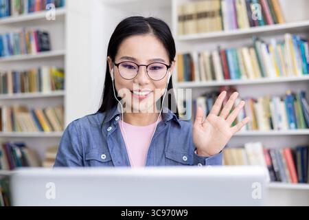Eine Frau winkt während eines Videoanrufs mit Brille und Ohrhörern in einer Bibliothek mit Bücherregalen im Hintergrund. Stockfoto
