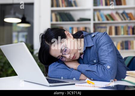 Ein müder Schüler schläft an einem Schreibtisch in einer Bibliothek, mit einem Laptop und Büchern in der Nähe. Stockfoto