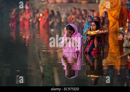 Bogura, Bangladesch - 20. November 2023: Blick auf Frauen in farbenfrohen Saris, die in ruhigen Gewässern stehen und Opfer halten, während ihre Reflexionen gegen die dunkle Flussoberfläche schimmern. Stockfoto