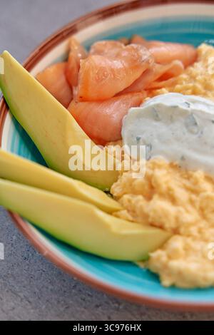 Ein farbenfroher Teller mit cremigen Rührei, Avocado-Scheiben, herzhaftem geräucherten Lachs und einer Kräuterjoghurtsauce für ein köstliches und nahrhaftes Ich Stockfoto