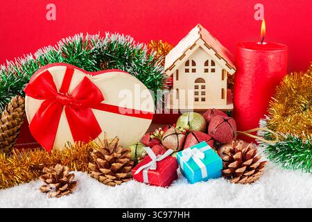 Weihnachts- oder Neujahrskarte. Brennende rote Kerze, Kegel, Geschenkboxen, dekoratives Haus, Spielzeug und Spaghetti vor rotem Hintergrund Stockfoto