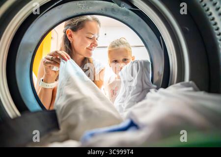 Mutter und Tochter waschen gemeinsam in einem öffentlichen Selbstbedienungswaschsalon, teilen sich die Hausaufgaben, bauen Verantwortung auf und arbeiten zusammen Stockfoto